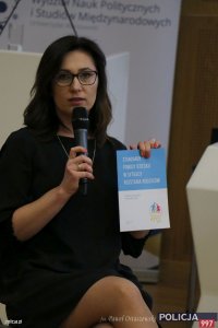 kobieta siedząca na krześle pokazuje ulotkę podczas ogólnopolskiej konferencji eksperckiej pn. „System Poszukiwań Osób Zaginionych - wyzwania XXI wieku” w związku z Międzynarodowym Dniem Dziecka Zaginionego