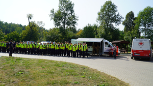 grupa policjantów stoi w dwuszeregu, obok pojazdy służb ratunkowych