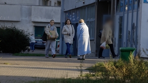 Zdezorientowana, zagubiona kobieta ubrana w jasnoniebieski szlafrok i kapcie stoi przed budynkiem. W jej kierunku idą dwie młode kobiety.